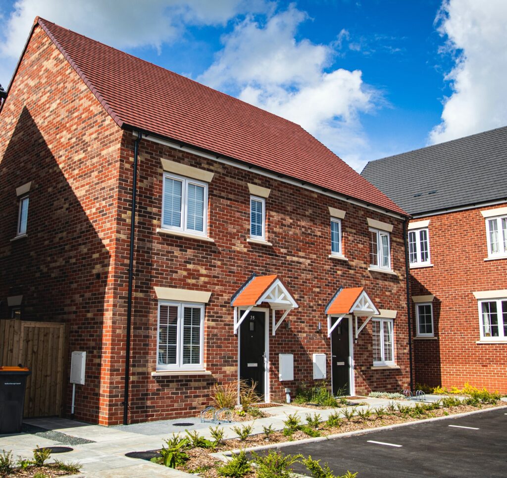 Photograph of semi-detached, red brick modern houses in a UK street. There are parking spaces outside in the foreground.